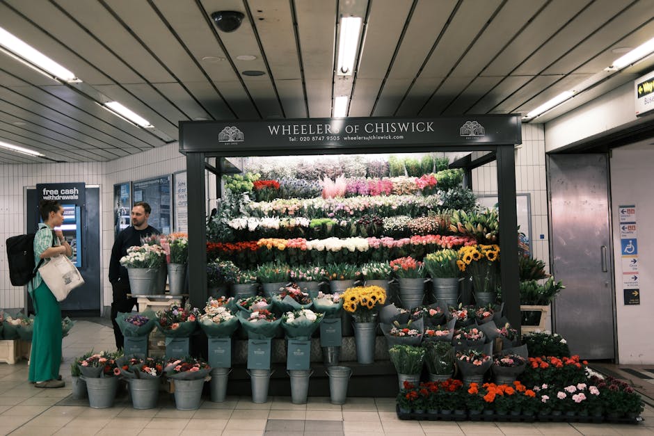 A vibrant display of fresh flowers arranged in a floral stand at Arsenal Station, featuring various types of blooms including pink roses, white carnations, yellow sunflowers, purple irises, and red tu