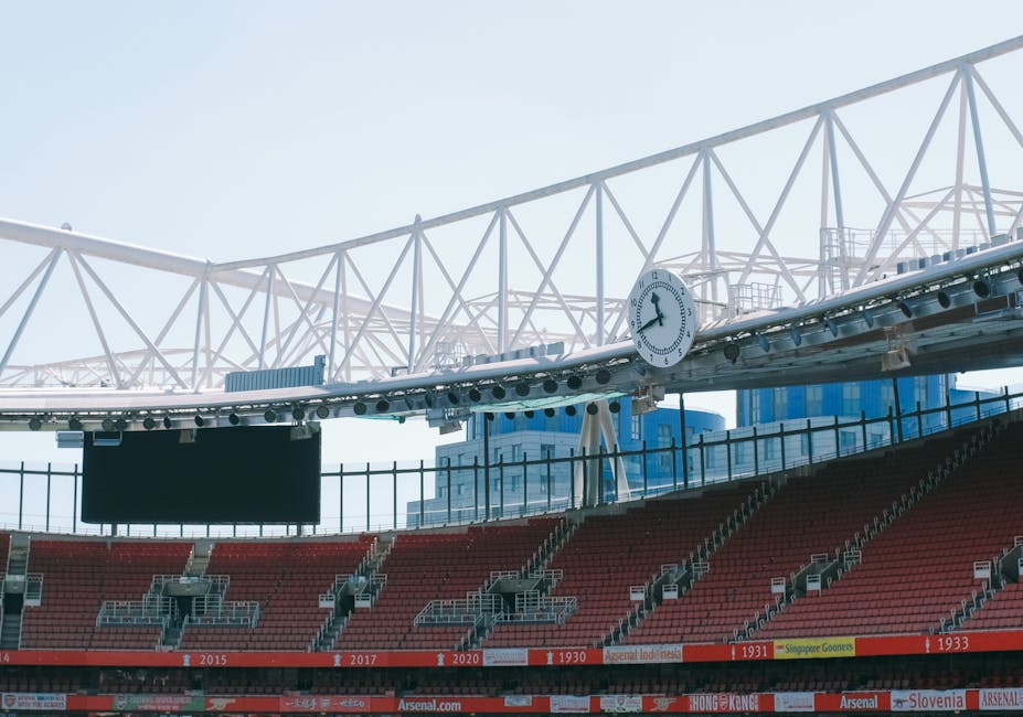 A section of the Emirates Stadium in Highbury, featuring red stadium seats, a white architectural roof structure with crisscrossing beams, and a round clock displaying approximately 10:07. The scene i