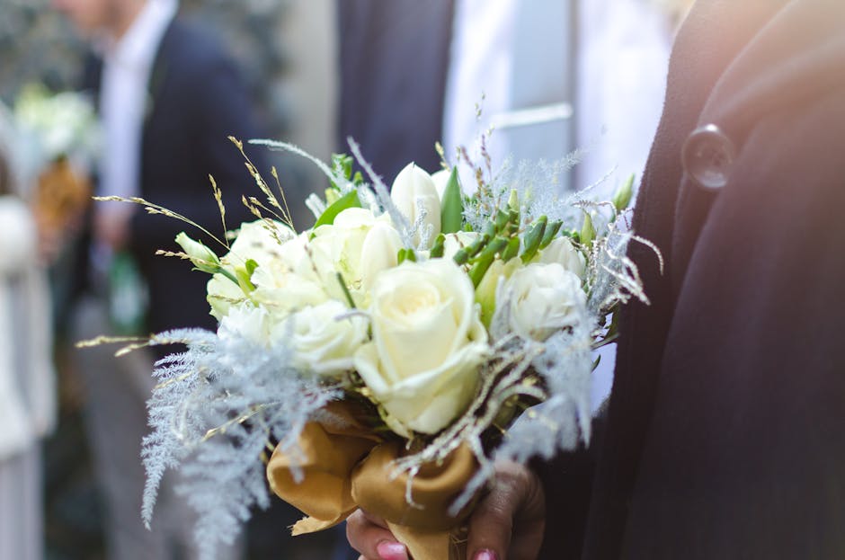 A close-up of a bridal bouquet featuring creamy white roses, delicate white lisianthus, and sprigs of dusty miller foliage, arranged in a natural, rounded style. The bouquet is wrapped with a gold sat