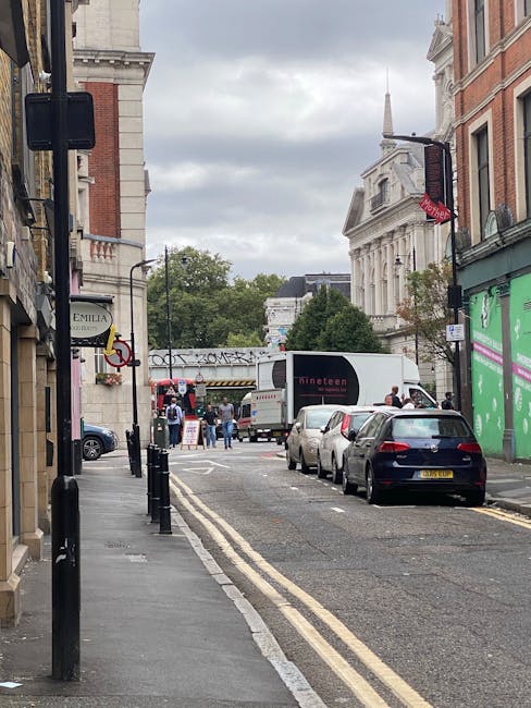 Image of a narrow street near Arsenal Station showing parked cars along the curb, with a delivery truck and pedestrians in the background. The scene includes classic London architecture with red brick