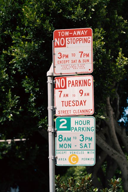 A close-up photograph of a vertical street sign pole with multiple signs against a backdrop of lush green trees and a clear blue sky. The top sign is red and white, indicating 'TOW-AWAY NO STOPPING 3P
