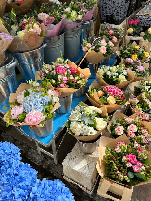 A display of various floral arrangements and bouquets featuring pink and white roses, hydrangeas, and other pastel-colored flowers, arranged in metal buckets and wrapped floral paper. The bouquets are