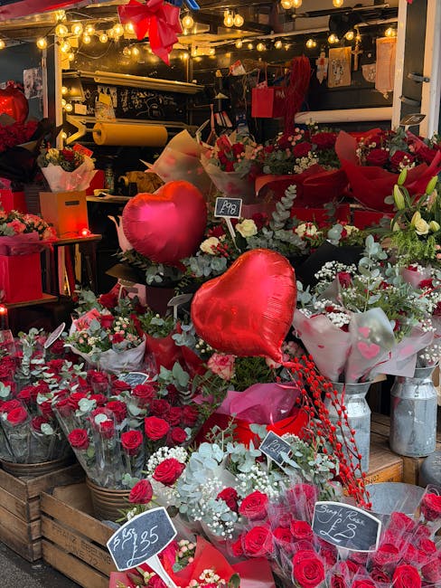 A vibrant display of fresh floral arrangements and bouquets featuring red roses, white blooms, and greenery, arranged on wooden crates and tables inside a florist shop. Bright red heart-shaped balloon
