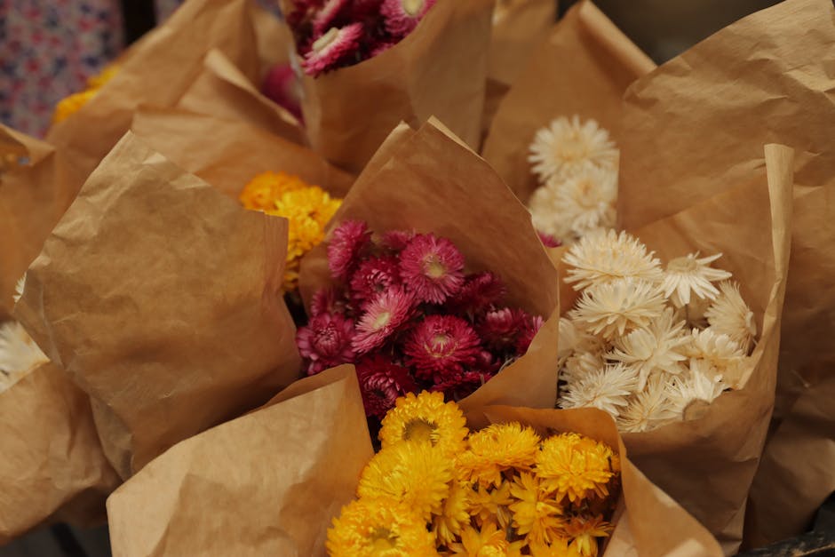 A collection of fresh flower bouquets wrapped in light brown kraft paper, arranged on a surface. The bouquets feature vibrant pink chrysanthemums, bright yellow daisies, and white spider chrysanthemum