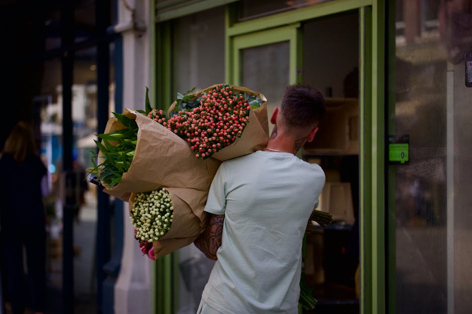 A person dressed in a light-colored T-shirt is seen from behind, carrying a large bouquet of fresh flowers wrapped in brown paper. The bouquet features vibrant pink and white varieties, with green fol
