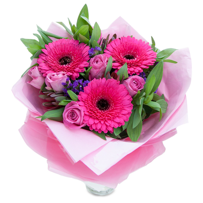 Playful yet elegant, this bouquet balances boldness and tenderness through a palette of fuchsia, lavender-pink and soft green. Three large Gerbera daisies dominate the view, their intense fuchsia petals fanning out around dark central discs that add striking contrast and focal drama. Around them, dusty-rose or lavender-pink roses sit as graceful companions, their petals tightly curled in fresh buds that promise to open into fuller blossoms. Small, papery clusters of purple statice provide a cool-toned punctuation across the arrangement, giving it an almost meadow-like texture against the smoother rose petals. A generous base of greenery-broad salal leaves and sprigs of aromatic eucalyptus-wraps the flowers in a lush, living backdrop that enhances the pinks and offers subtle scent when handled. The bouquet's presentation is thoughtful: an inner layer of soft, matte pale-pink tissue forms a gentle cradle for the stems, while an outer wrap of sheer, crinkled fuchsia-pink cellophane adds a slight iridescent sheen and keeps the composition cohesive. The top rim of a clear glass vase peeks through at the bottom, indicating the flowers are arranged for immediate display. I can easily picture this bright, tender arrangement brightening an apartment on Drayton Park or being carried home from a walk through Highbury Fields-perfect for celebrating a birthday, marking an anniversary, or saying 'thank you' with warmth and care.